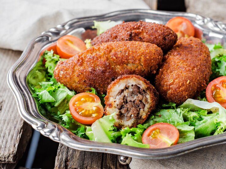 Schräge Frontalsicht: Auf einem runden silbernen Teller liegen Kibbeh, orienatlische Bulgur-Bällchen, die mit Hackfleisch gefüllt sind. Drum sind grüne Salatblätter und halbierte roten Tomaten.