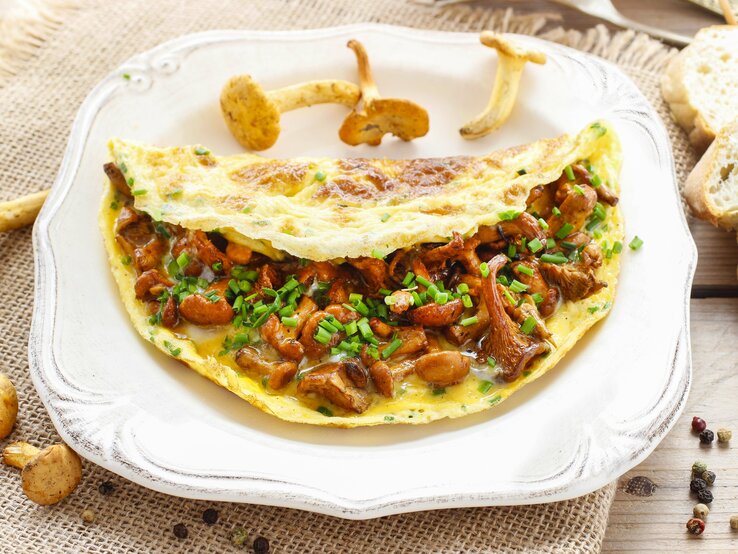 Auf einem weißen Teller liegt ein Pfifferling-Omelette. Um den Teller herum liegen frische Pfifferlinge, Pfefferkörnern und Weißbrotscheiben. | © Shutterstock / Agnes Kantaruk
