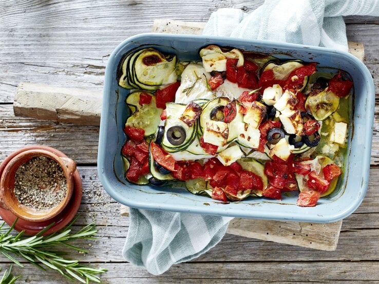 Mediterraner Fischauflauf in einer blauen Auflaufform. Daneben liegen Gewürze, Das Ganze auf einem hölzernen Untergrund. 