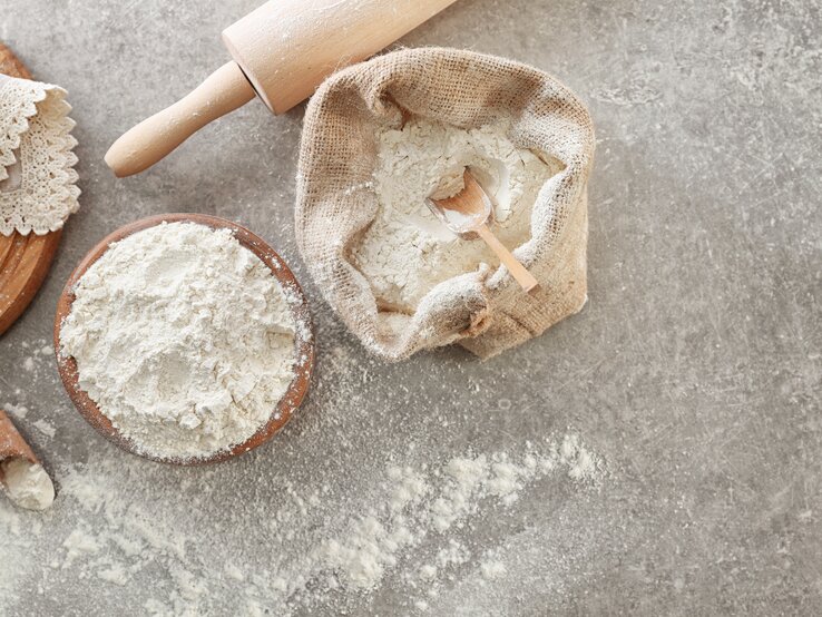 Draufsicht: Auf einem grauen Untergrund stehen ein Leinensack und eine Schüssel mit Self Rising Flour. Daneben liegen eine Backrolle sowie ein Brettchen mit einer Spitzendecke.