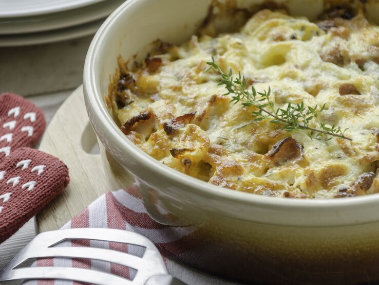 Kretanische Kartoffeln in einer hellen Auflaufform mit frischem Kraut neben Ofenhandschuhen und einem Pfannenwender auf einem hellen Holzbrett neben weiteren Tellern auf einem hellen Tisch. 