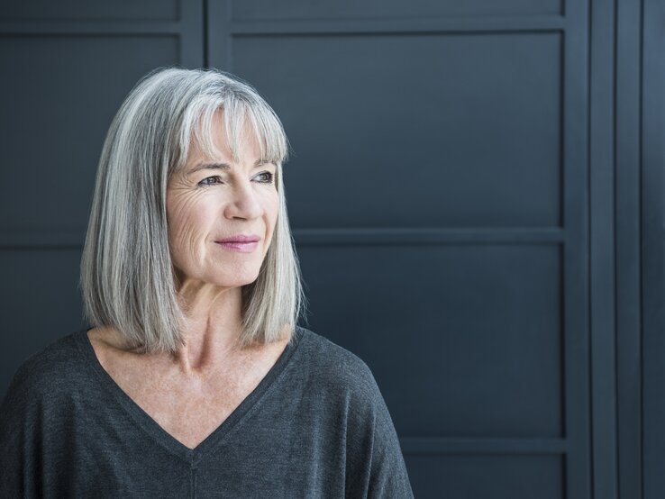 graue-Haare Eine Frau mit grauen Haaren und einem dunkelgrauen Oberteil blickt nachdenklich zur Seite, vor einem dunkelblauen Hintergrund.