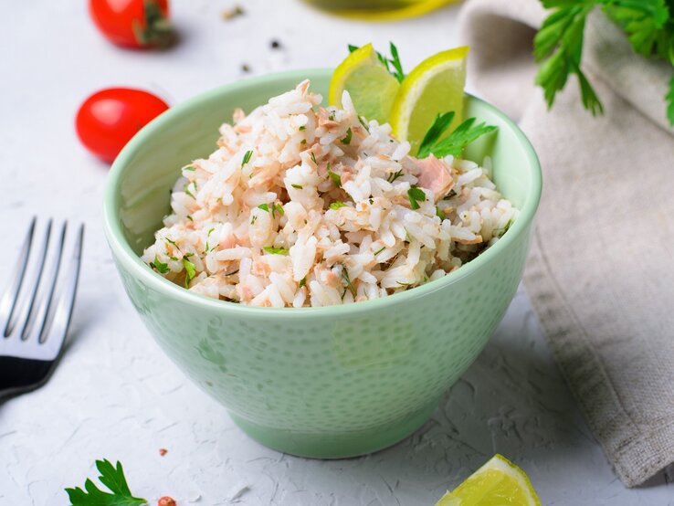 Eine kleine grüne Schale mit dem Thunfisch-Reis auf einem grauen Tisch mit mausgrauer Decke. Daneben Gabel, Kirschtomaten und frische Petersilie.