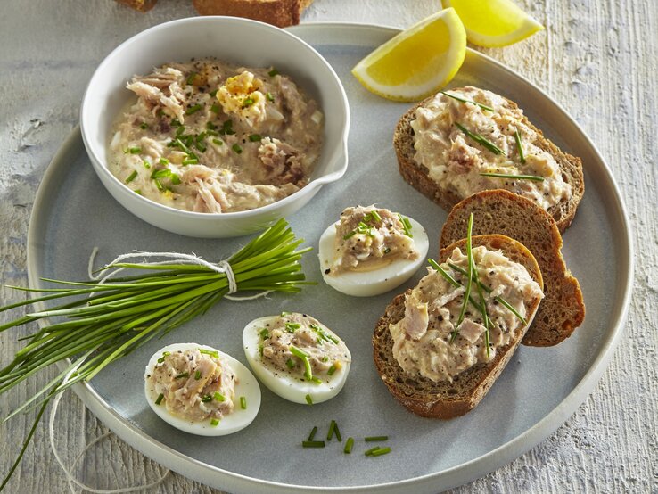Thunfisch-Ei-Aufstrich in einer Schale und auf Brot und Ei. Daneben liegt Schnittlauch und geschnittene Zitrone. 