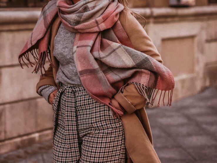 Eine Frau trägt angesagte Strickmode für den Herbst, wie eine karrierte Hose, einen graumelierten Pullover und einen XXL-Schal in braun, rosa und weiß. Dazu kombiniert sie einen hellbraunen Mantel während sie durch die Straßen läuft.