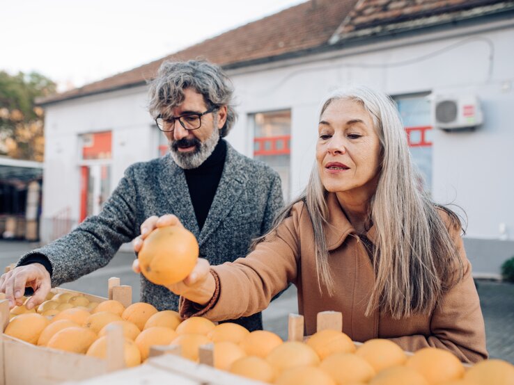  Ein älteres Paar mit grauen Haaren steht an einem Obststand und wählt gemeinsam frische Orangen aus. Der Mann, der eine Brille und einen grauen Wollmantel trägt, hält eine Orange hoch, während die Frau mit langen, glatten Haaren und einem braunen Mantel aufmerksam zusieht. Sie scheinen die Qualität der Früchte zu begutachten, was auf ein Bewusstsein für gesunde Ernährung hinweist. Im Hintergrund ist ein Teil eines Gebäudes mit einem roten Fensterrahmen zu sehen, was auf ein öffentliches Umfeld wie einen Markt hinweisen könnte.