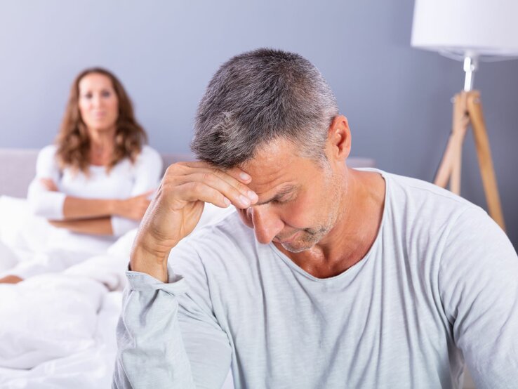 Paar sitzt enttäuscht am Bettrand Eine Frau liegt im Bett, ihr Partner sitzt geknickt am Bettrand und schaut nach unten.