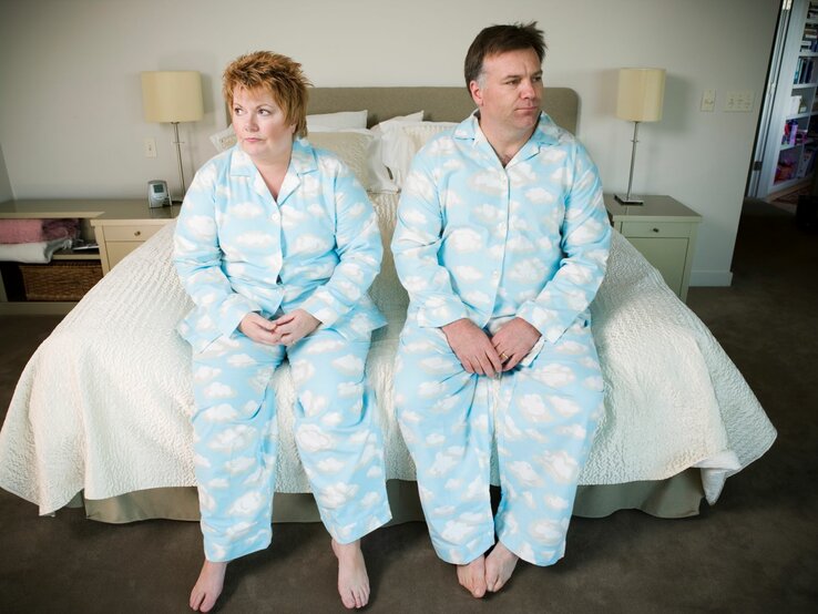 Ein reiferes Paar sitzt im Pyjama nebeneinander auf dem Bettrand und schaut etwas frustriert in die jeweils andere Richtung | © Getty Images / Big Cheese Photo