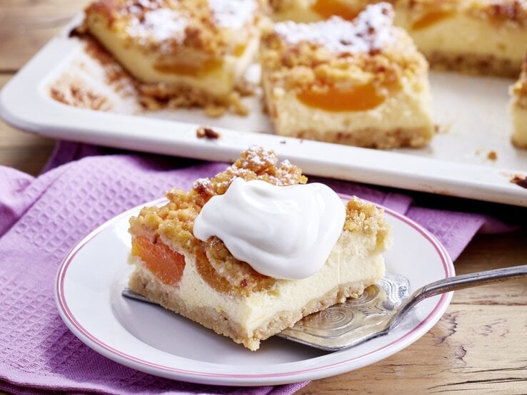 Unser Aprikosen-Schmand-Kuchen ist super saftig und fruchtig. Mit einem Klecks Schlagsahne schmeckt er noch besser! Auf einem Teller ist ein Stück Aprikosen-Schmand-Kuchen serviert. Darauf ist ein Klecks Schlagsahne.