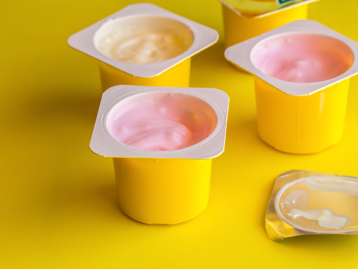 Fruchtaromatisierter Jogurt in gelben Plastikbechern auf hellgelbem Hintergrund mit silbernem Foliendeckel - Yogurt-Cup-Hintergrund mit selektivem Fokus