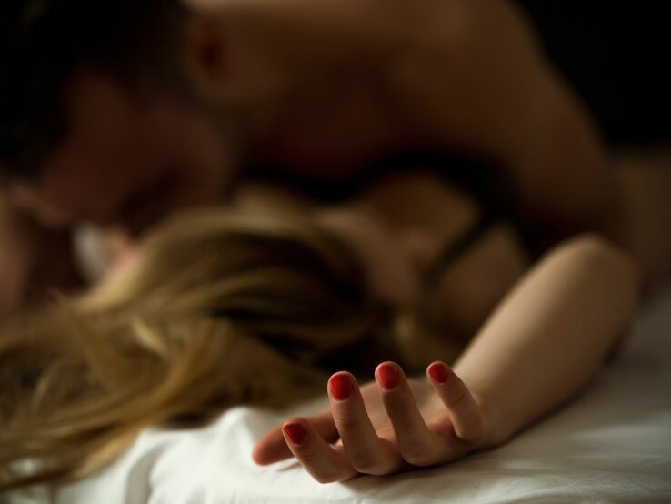 Orgasmus Tricks Frau Ein unscharfes Bild zeigt zwei Personen in einem intimen Moment auf einem Bett. Der dunkelhaarige Mann scheint die blonde Frau am Nacken zu küssen. Der Fokus liegt auf einer Hand mit rot lackierten Nägeln, die sich auf einer weißen Bettdecke abzeichnet, was auf eine starke Emotion hindeutet.