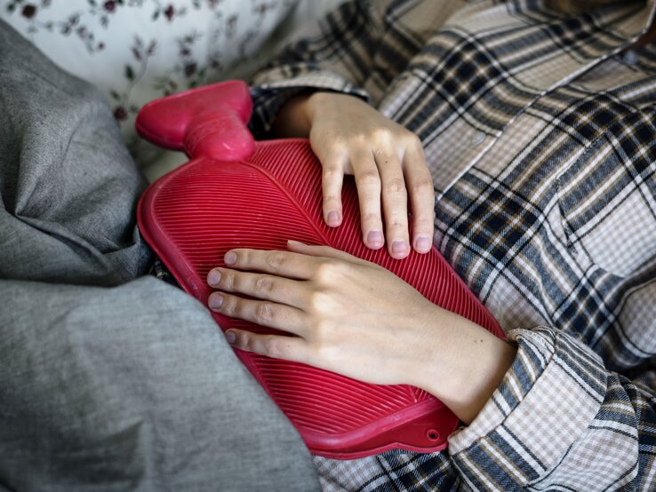  Das Bild zeigt eine Person, die entspannt auf einer Couch oder einem Bett liegt, mit einer rot gefärbten Wärmflasche auf ihrem Bauch. Die Person trägt eine karierte Jacke und liegt unter einer grauen Decke. Ihre Hände liegen sanft auf der Wärmflasche, was darauf hindeuten könnte, dass sie Wärme und Komfort sucht, möglicherweise um Schmerzen oder Unbehagen zu lindern. Das Muster der Jacke und die Textur der Wärmflasche sind deutlich sichtbar und erzeugen einen gemütlichen und behaglichen Eindruck.