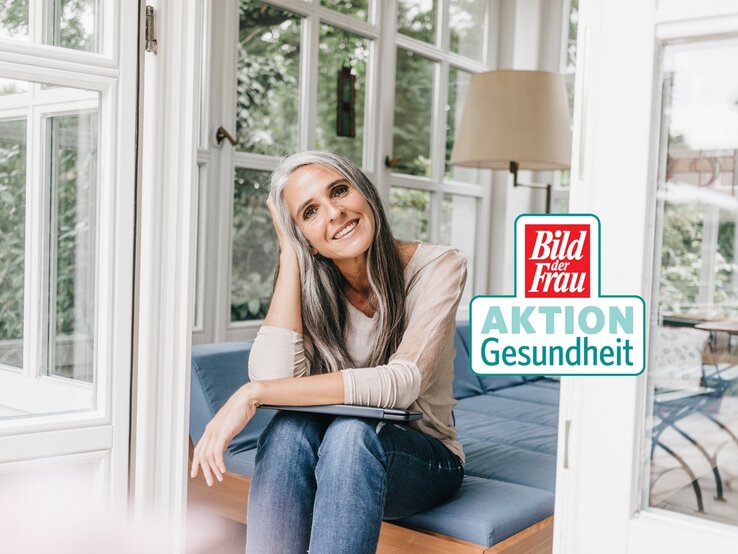 Eine Frau mittleren Alters mit graumelierten Haaren und einem warmen Lächeln sitzt entspannt auf einer blauen Couch vor einem großen Fenster. Sie trägt ein lässiges, beiges Langarmshirt und dunkelblaue Jeans. Das Zimmer ist hell und freundlich, mit einem Blick auf grüne Pflanzen außerhalb des Fensters. Auf dem Bild ist auch ein Logo "Bild der Frau AKTION Gesundheit" erkennbar.