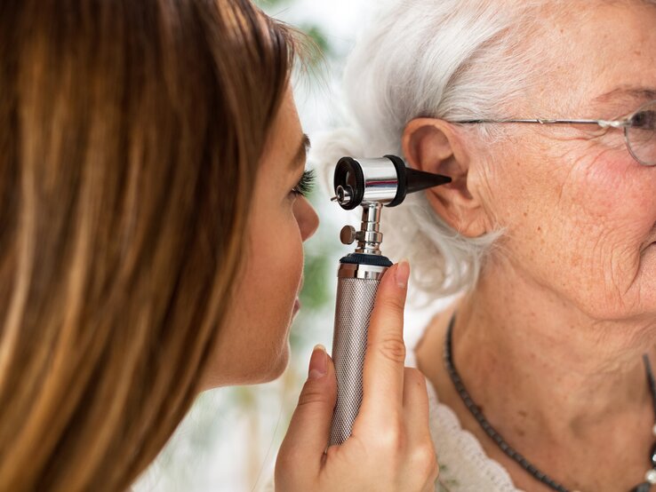 Eine junge Ohrenärztin schaut einer älteren Patientin mit einem Instrument ins Ohr, um zu prüfen, ob sich Eiter gesammelt hat.