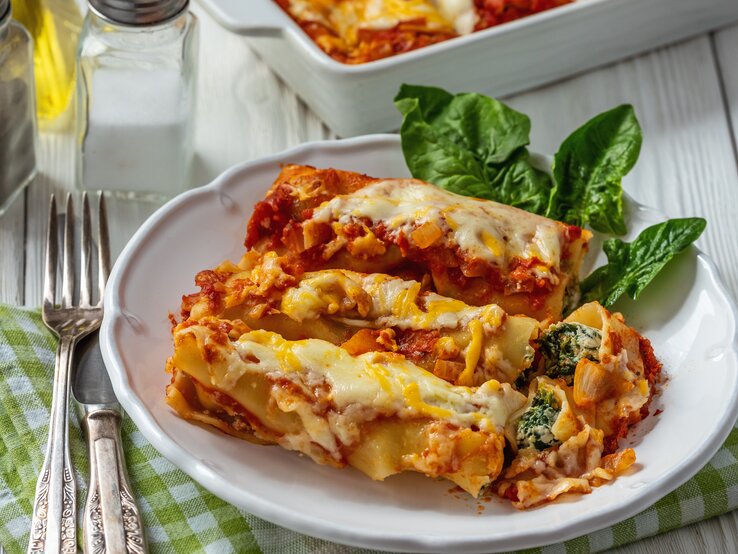 Weißer Teller belegt mit Cannelloni mit Ricotta-Spinat-Füllung und Tomatensoße auf hölzernem Untergrund.