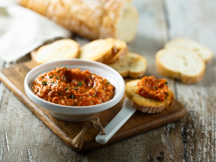 Eine Schale Tomate-Walnuss-Aufstrich auf einem Brett, daneben Baguettescheiben.