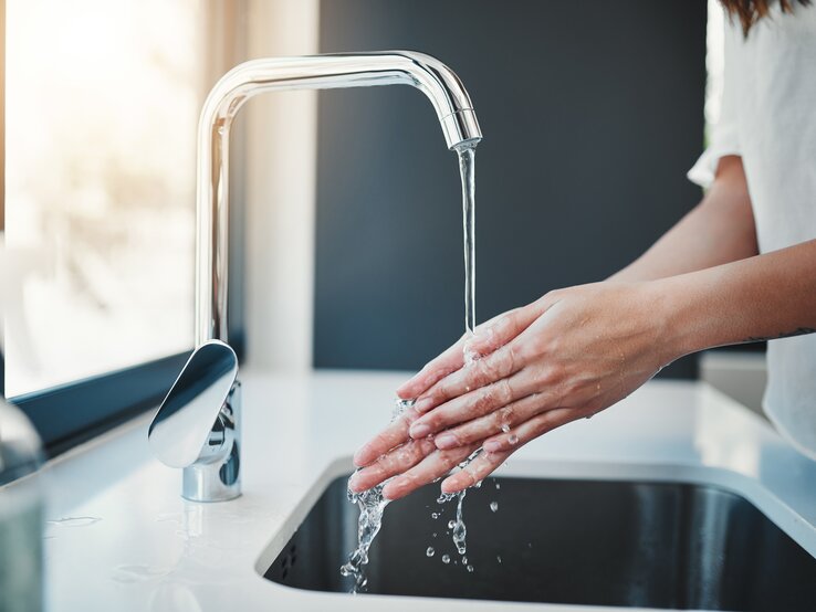 Eine Person wäscht sich sorgfältig die Hände mit Wasser aus einem modernen Wasserhahn über einem Küchenspülbecken.