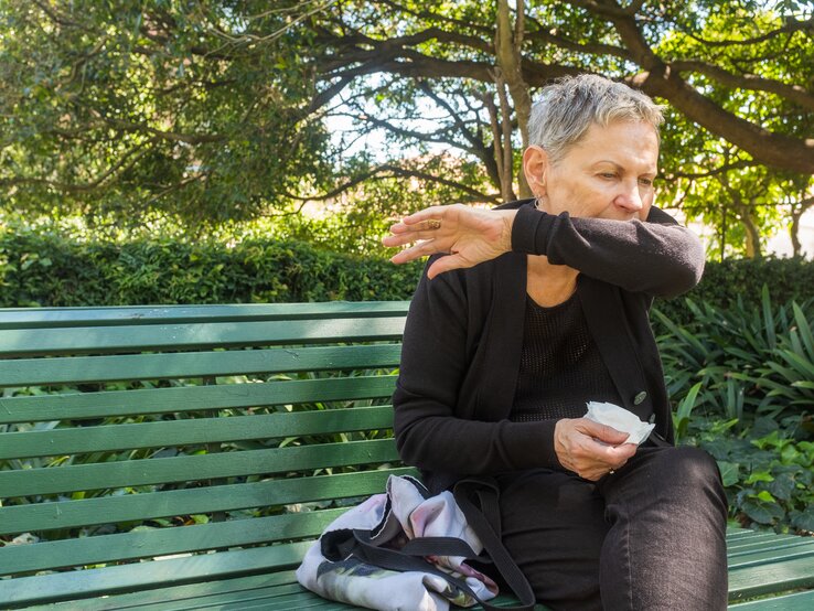 Eine ältere Frau mit kurzen grauen Haaren, die eine schwarze Jeans und ein schwarzes Oberteil trägt, sitzt auf einer grünen Parkbank im Freien. Sie hält ein Taschentuch in der rechten Hand und hustet in ihre Armbeuge.