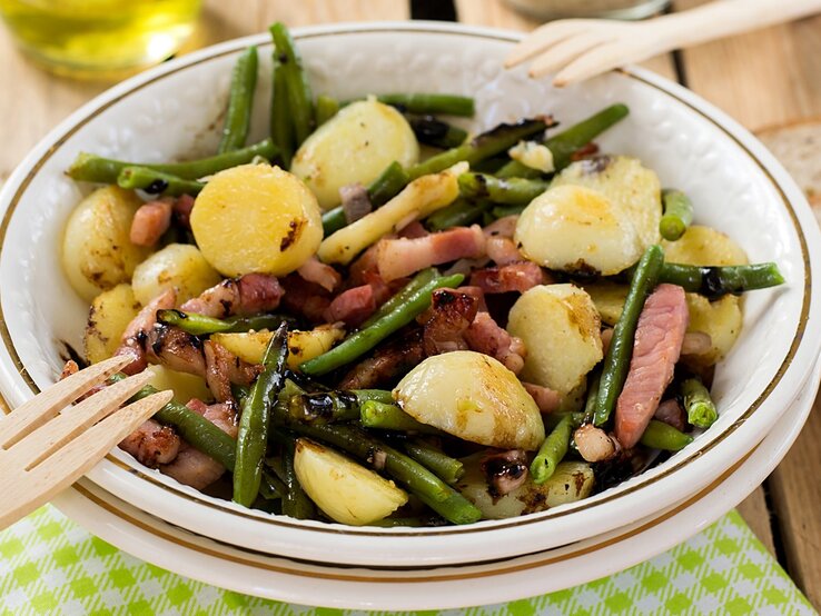 Der Kartoffelsalat auf einem weißen Teller. Alles auf einem rustikalen Holztisch mit weiß-grün-karierter Decke von schräg oben fotografiert.