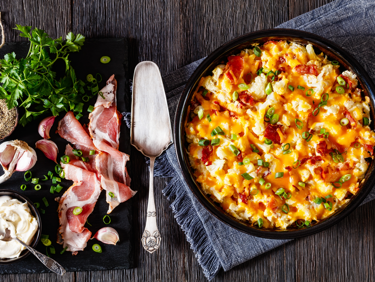 Blumenkohlauflauf mit Speck in schwarzer Form auf dunklem Holz. Daneben liegen Frühlingszwiebeln, Speck und Knoblauch. 