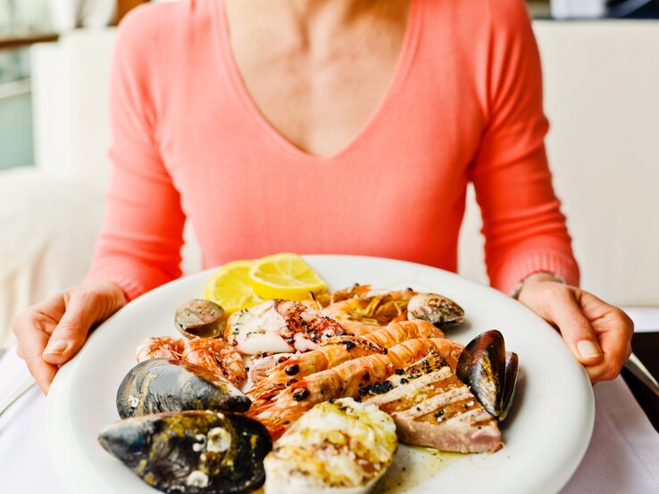 Eine Person im rosa Oberteil hält einen Teller mit Garnelen, Muscheln, Tintenfisch und Zitronenscheiben.