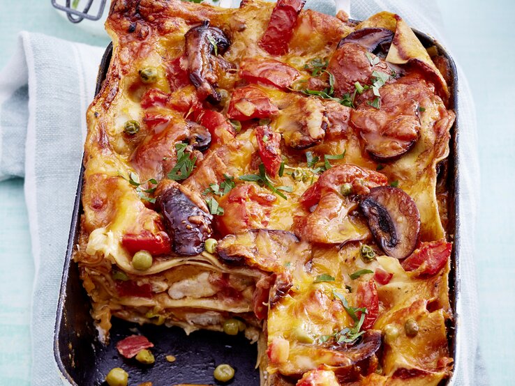 Hähnchen-Lasagne mit Pilzen und frischen Tomaten, angeschnitten in einer großen Auflaufform auf einem weißen Tuch serviert. 