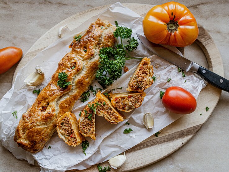 Hackfleisch-Blätterteig-Rolle auf einem Holzbrett. Daneben ein Messer und mehrere Tomaten. 