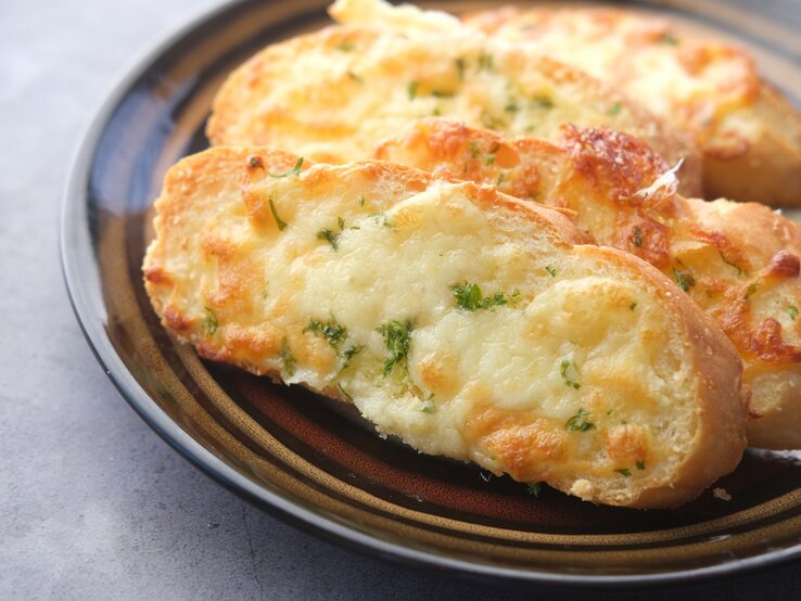 Käse-Knoblauch-Brot auf einem gemusterten Teller. Das Ganze auf einem grauen Untergrund. 