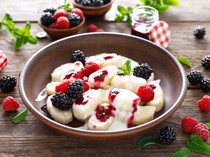 Brauner Teller gefüllt mit süßen Gnocchi, Sahne und Beeren. Darum herum liegen auf einer Holzplatte Beeren und Minze. 