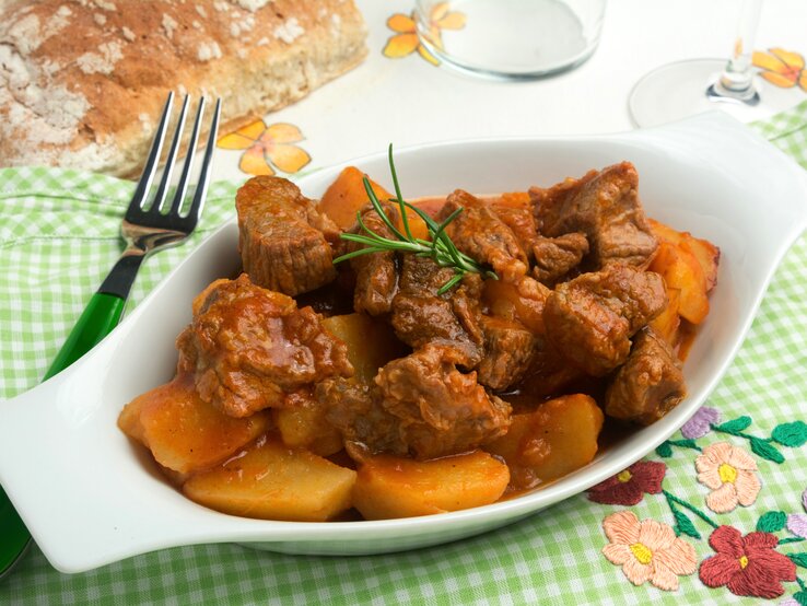 Draufsicht schräg: Auf einem grün-weiß-kariertem Tuch ist eine kleine Schale mit Spezzatino di manzo, ein italienisches Gulasch. Drum herum liegen eine Gabel und ein Stück Weißbrot.