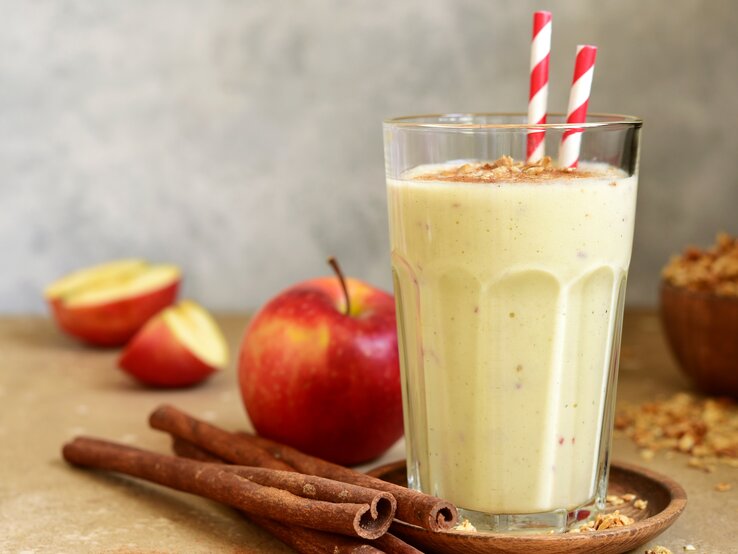 Ein Glas mit dem Smoothie auf einem hellbraunem Untergrund. Daneben ein paar Zimtstangen und ein paar Apfelspalten.