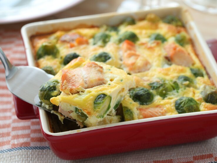 Ein Rosenkohl-Lachs-Gratin in einer weiß-roten Form steht auf einem gemusterten Geschirrtuch. Das Gratin ist bereits angeschnitten und ein Stück wird mit einem Tortenheber herausgehoben.