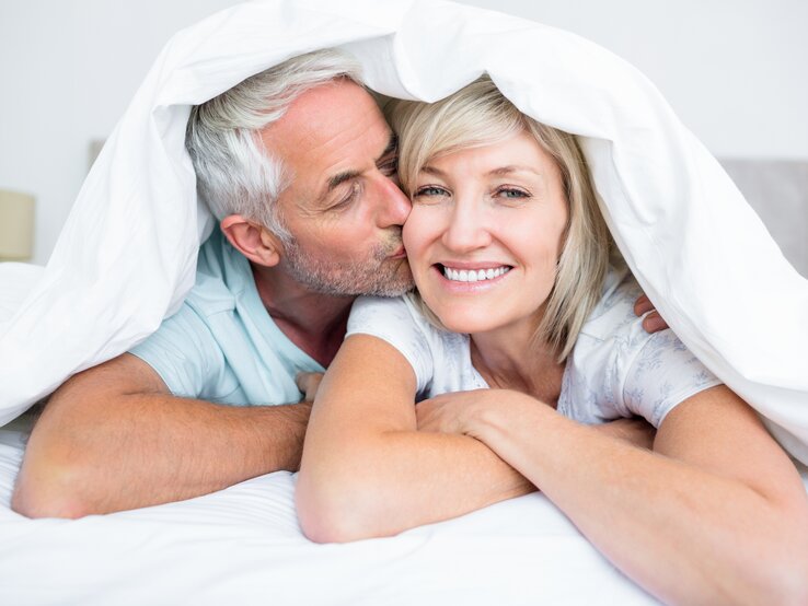 Älteres Ehepaar liegt im Bett mit der Bettdecke über den Köpfen, er küsst sie auf die Wange, sie lacht