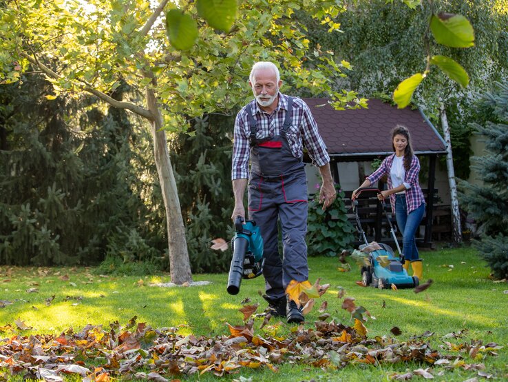 Ein älterer Mann mit Laubbläser, und im Hintergrund eine junge Frau mit einem Rasenmäher, arbeiten in einem Garten.