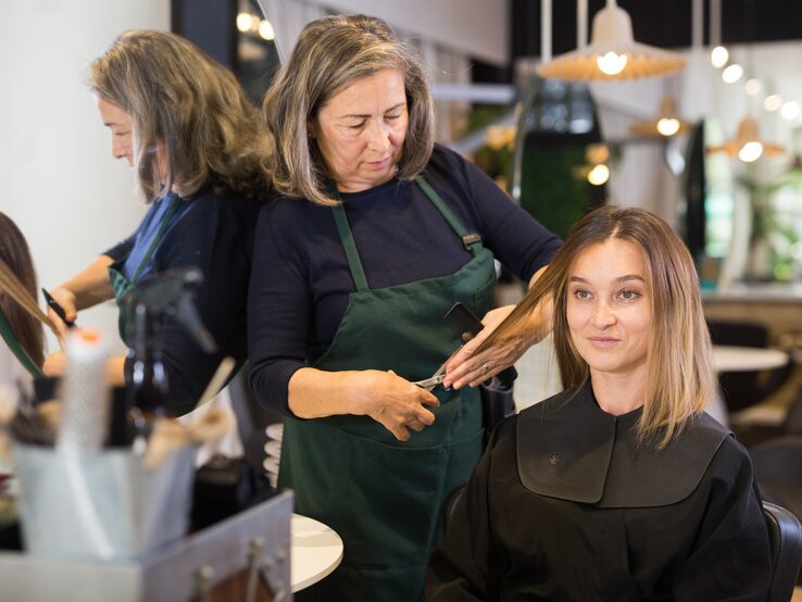 Eine ältere Friseurin mit grauen Haaren schneidet ihrer jüngeren Kundin, die blonde Haare hat und auf dem Friseurstuhl sitzt, die Haare. Vielleicht bekommt Sie eine der Trendfrisuren im Herbst 2023 geschnitten.