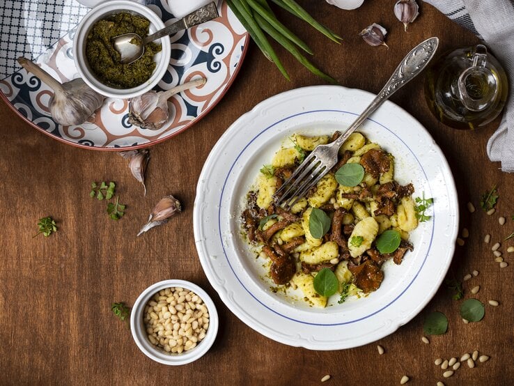 Gnocchi-Pfifferling-Pfanne mit Pesto und Parmesan auf weißem Teller. Dieser steht auf einer braunen Oberfläche, umringt von verschiedenen Zutaten. 