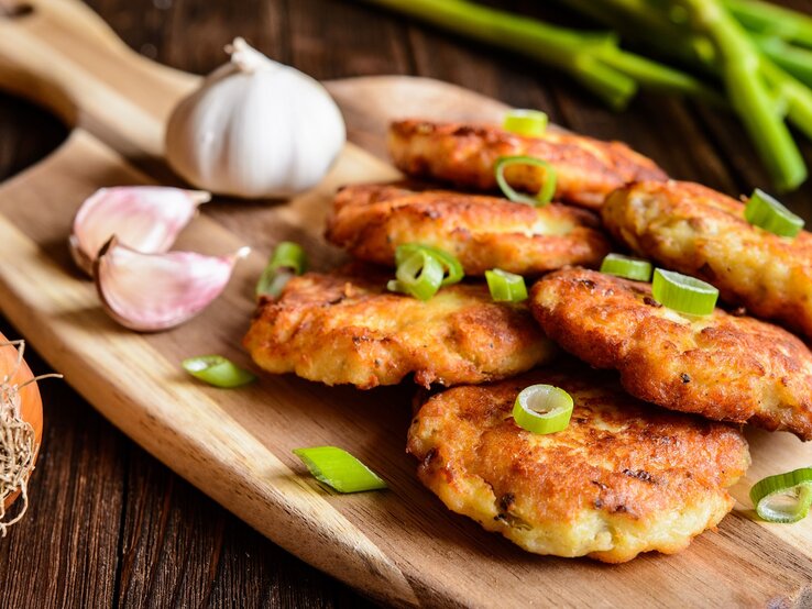 Mehrere Thunfischbratlinge auf einem Holzbrett und einem rustikalen Holztisch mit Knoblauch und Frühlingszwiebel im Hintergrund.