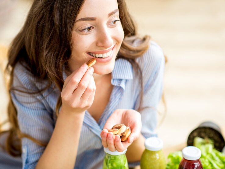 Eine junge Frau mit langen braunen Haaren und einem blau gestreiften Hemd sitzt lächelnd an einem Tisch und isst Nüsse aus ihrer Handfläche. Vor ihr stehen frische grüne Smoothies und ein Salat, was auf eine gesunde Ernährung hindeutet. Sie scheint eine Pause zu genießen und wirkt entspannt und zufrieden in ihrer hellen, freundlichen Umgebung.