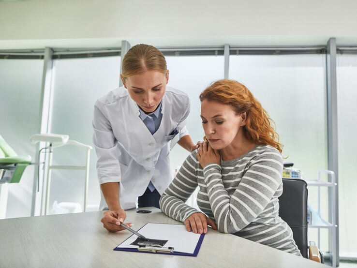 Eine Ärztin in weißem Kittel beugt sich über einen Tisch, um einer besorgten Patientin etwas auf einem Informationsblatt zu erklären. Die Patientin, erkennbar nachdenklich und vielleicht etwas besorgt, hört aufmerksam zu und stützt dabei ihr Kinn mit der Hand. Sie sitzen beide in einem hellen, klinischen Beratungszimmer.