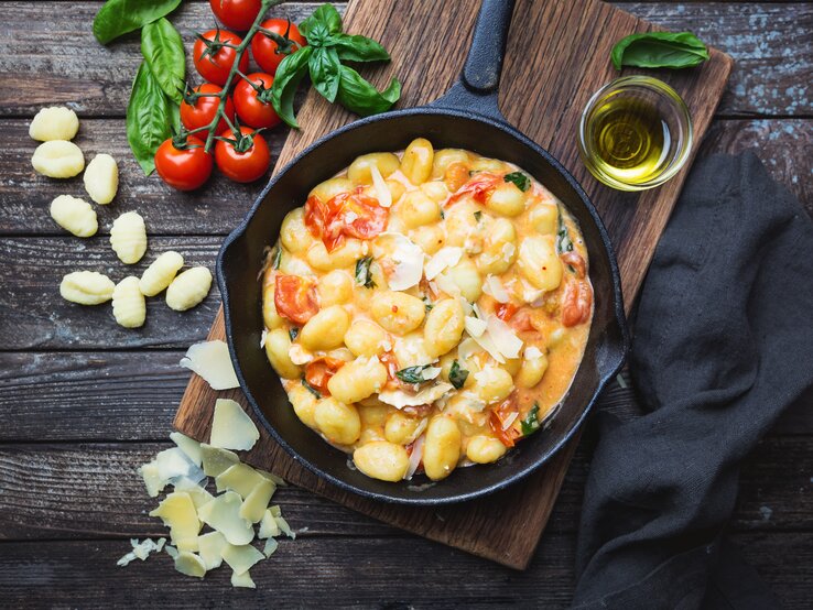 Gnocchi in Tomaten-Rahm-Soße in gusseiserner Pfanne auf Holztisch. Daneben liegen Tomaten und ungekochte Gnocchi.