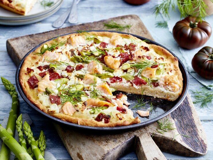 Angeschnittene Lachs-Feta-Pizza auf dunklem Teller. Drum herum Gemüse auf hellem Holzuntergrund. 