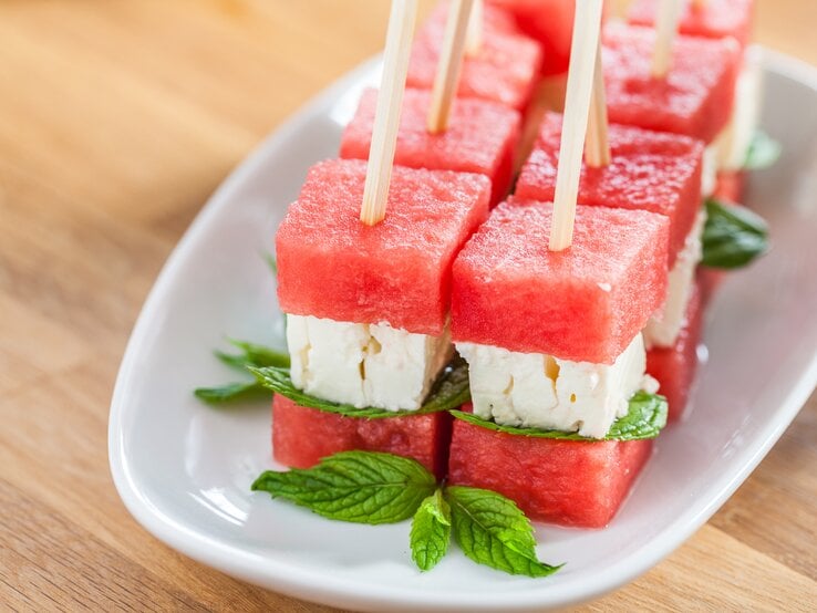 Wassermelonen-Feta-Spieße auf weißem Teller, angerichtet auf hölzernem Untergrund. 