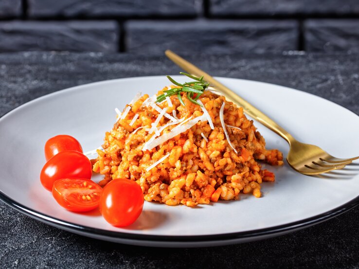 Schlotzig, tomatig und einfach! Ein Hackfleisch-Risotto ist perfekt für Risotto-Einsteiger! Hackfleisch-Risotto mit Cocktailtomaten auf weißem Teller mit Kupfergabel auf dunklem Untergrund. Draufsicht.