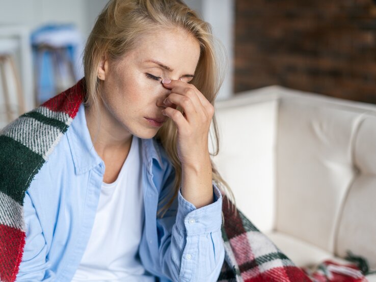 Zu sehen ist eine blonde Frau, die auf einem Sofa sitzt. Sie trägt ein hellblaues Hemd und darunter ein weißes Shirt. Sie hat eine Decke um ihre Schultern gelegt, und reibt mit der Hand zwischen ihren Augen, über der Nase. Die Augen sind geschlossen, die Frau scheint müde und erschöpft. 