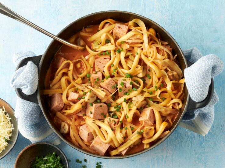 In einem Topf ist ein bayerischer Spätzle-Topf mit Leberkäse, Champignons und Kohl angerichtet. Daneben steht geriebener Käse.