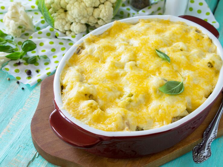 Draufsicht, schräg: Auf einem hellblauen Untergrund aus Holz steht eine ovale Ofenform mit einem Blumenkohl-Lachs-Auflauf mit Gouda auf einem Brettchen. Daneben liegt roher Blumenkohl.