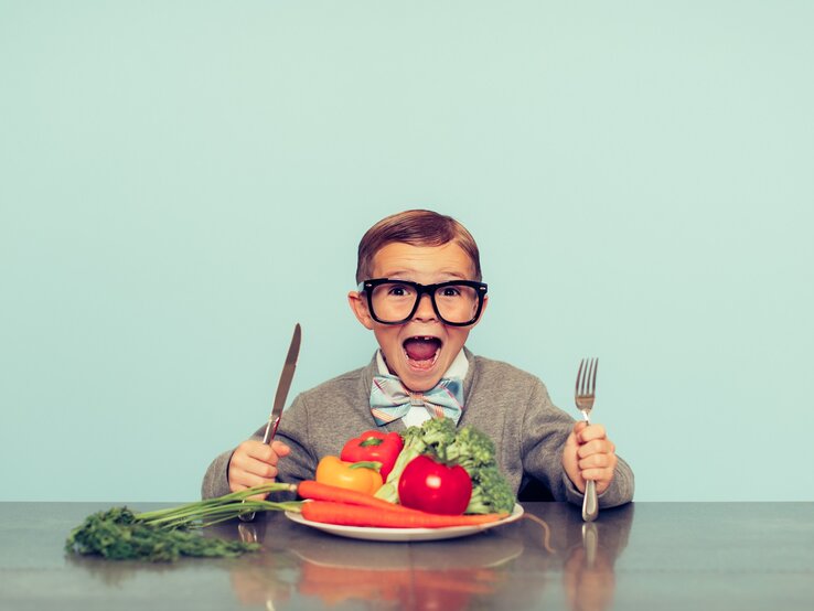 Vegane Ernährung bei Kindern: Ein Junge mit großer Brille und Fliege sitzt mit Besteck vor einem Teller mit Obst und Gemüse.