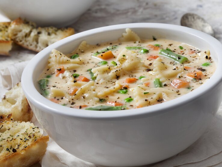 Suppenteller gefüllt mit Käse-Nudelsuppe mit Gemüse auf hellem Untergrund.