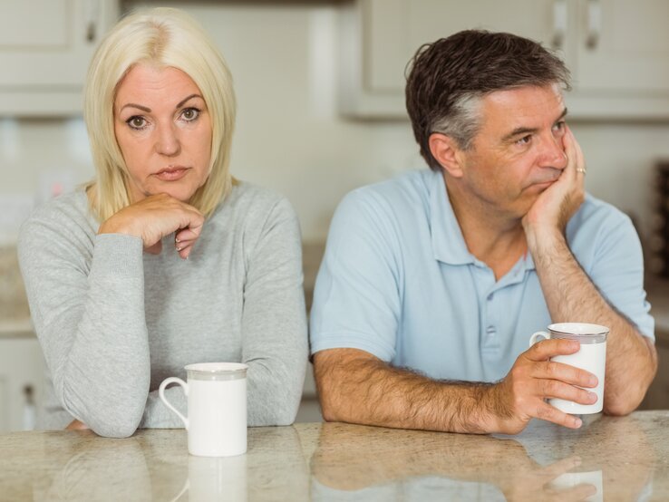 Ein Paar sitzt nebeneinander am Küchentisch, beide haben eine Kaffeetasse vor sich. Sie schaut traurig in die Kamera, er resigniert zu Seite.