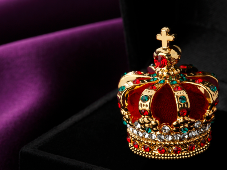 Ein prächtiges Modell einer Krone, verziert mit roten und blauen Edelsteinen sowie Diamantimitationen, ruht auf einer dunklen Samtoberfläche vor einem violett-schwarzen Hintergrund.